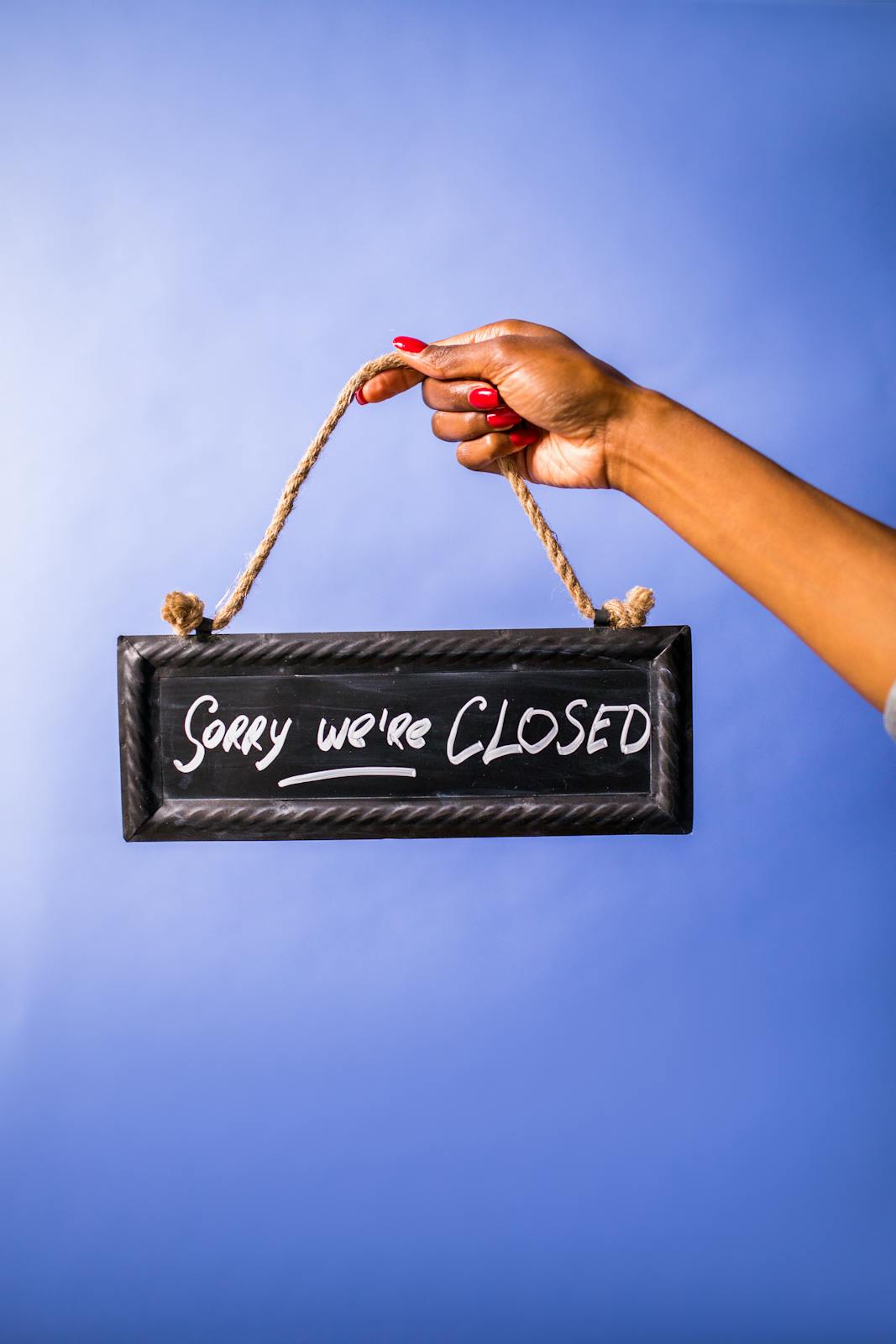 A woman's hand with red nails holds a 'Sorry We're Closed' sign against a blue background.